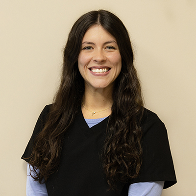 A smiling woman in a blue shirt, standing against a beige background, wearing a white lab coat and posing for the photograph.