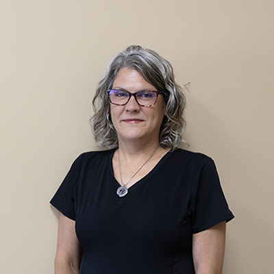 The image shows a woman standing against a wall, wearing glasses and a black top.