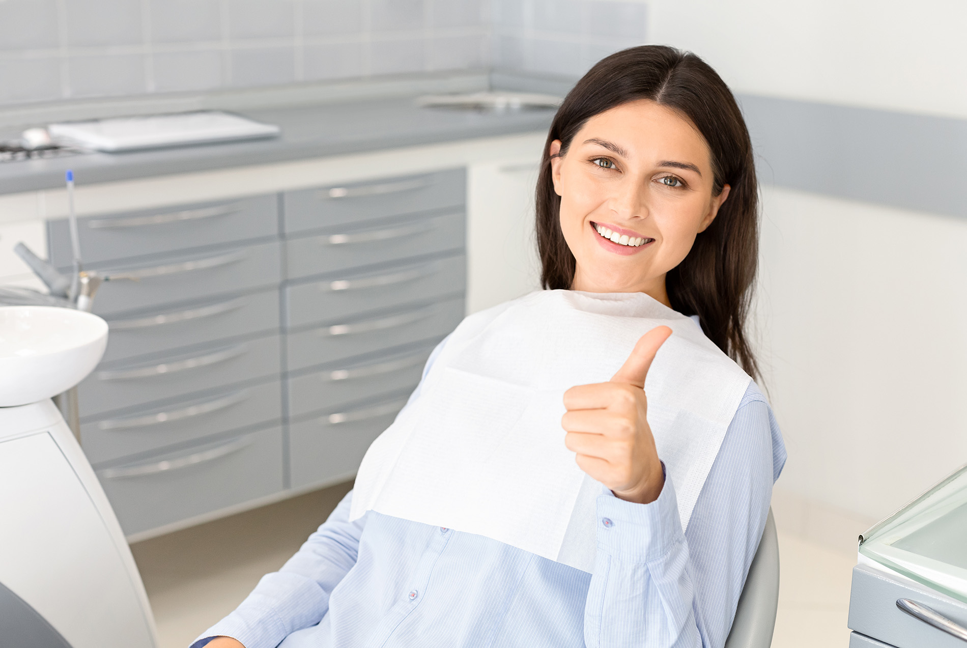 A woman with a thumbs-up gesture, sitting in a dental chair with a smile on her face.