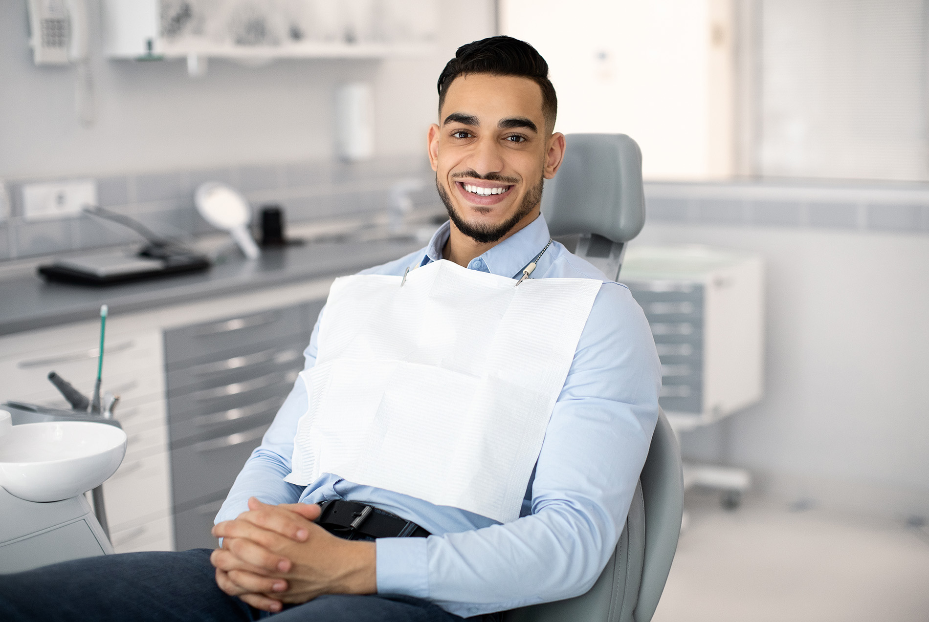 The image shows a man sitting in a dental chair, wearing a face mask and holding his chin with one hand. He is smiling and appears to be in a dental office setting, as indicated by the presence of dental equipment and a sink in the background.