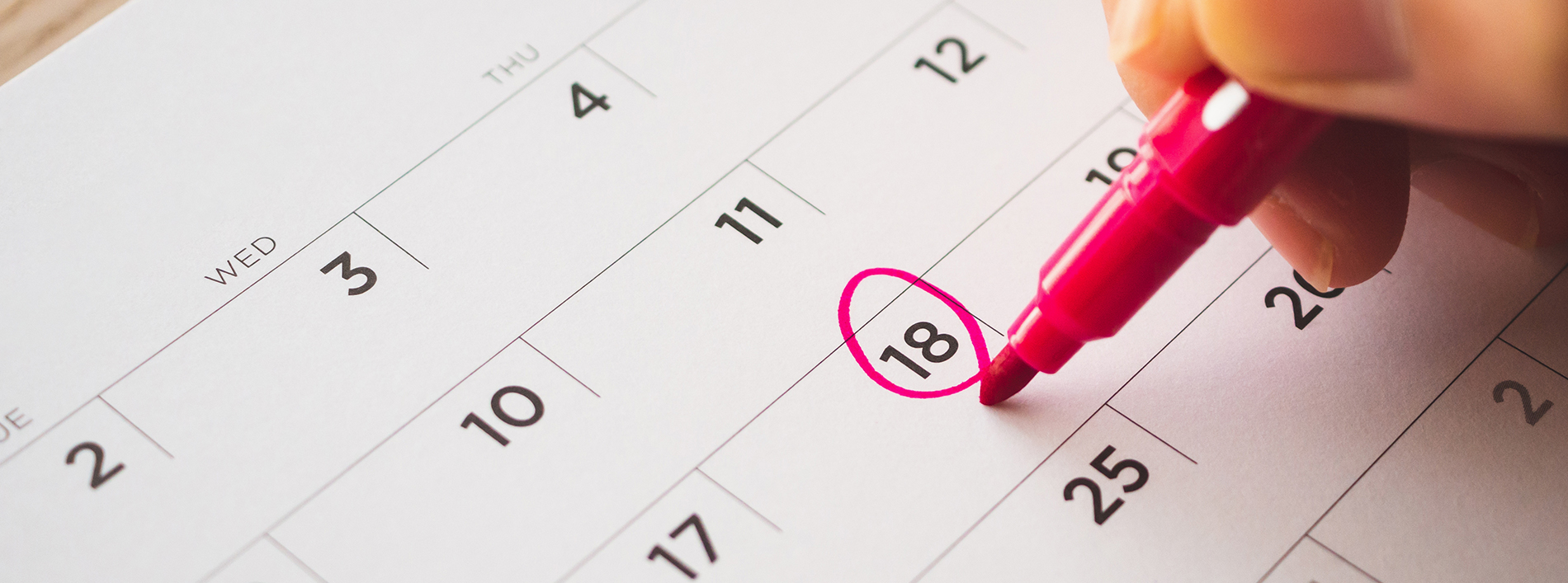 A close-up of a hand holding a pink marker, pointing to the 18th day on a calendar, with a blurred background.