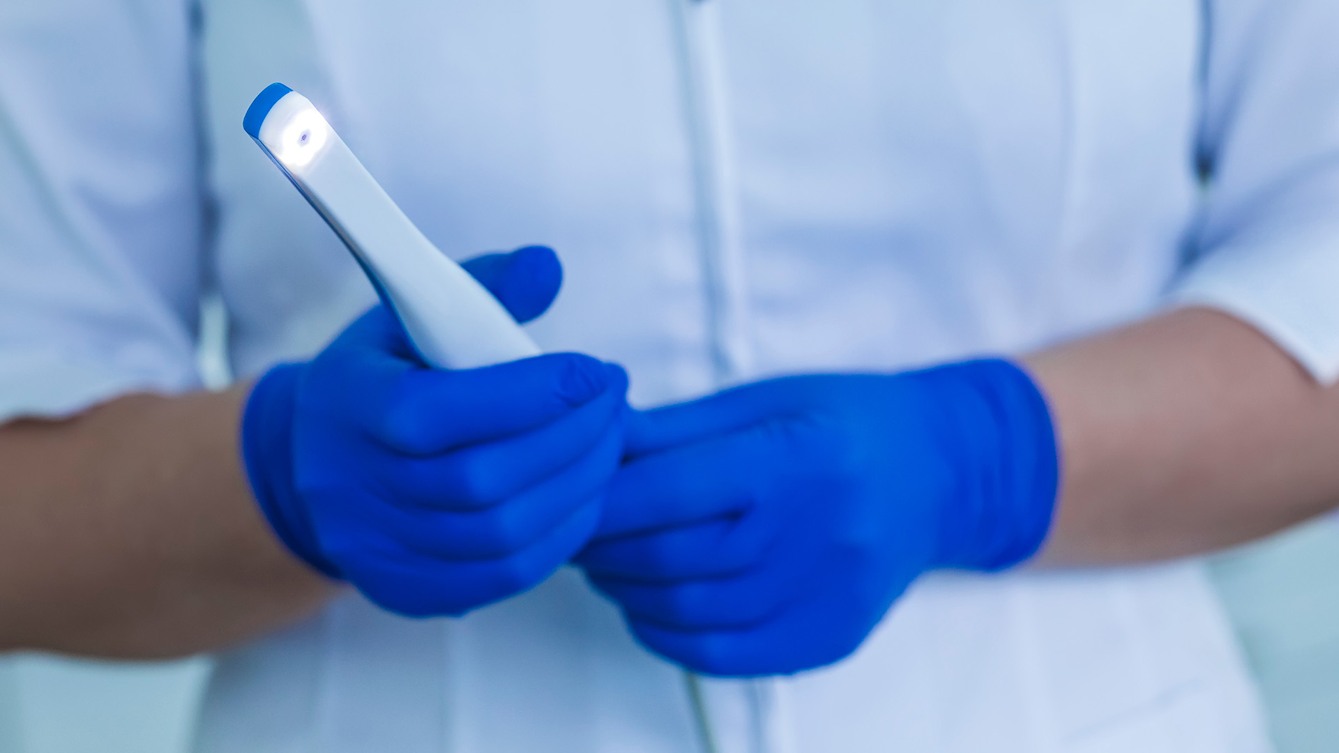 A healthcare worker in a blue gown and gloves is holding a smartphone with one hand, while the other hand is resting on their hip.