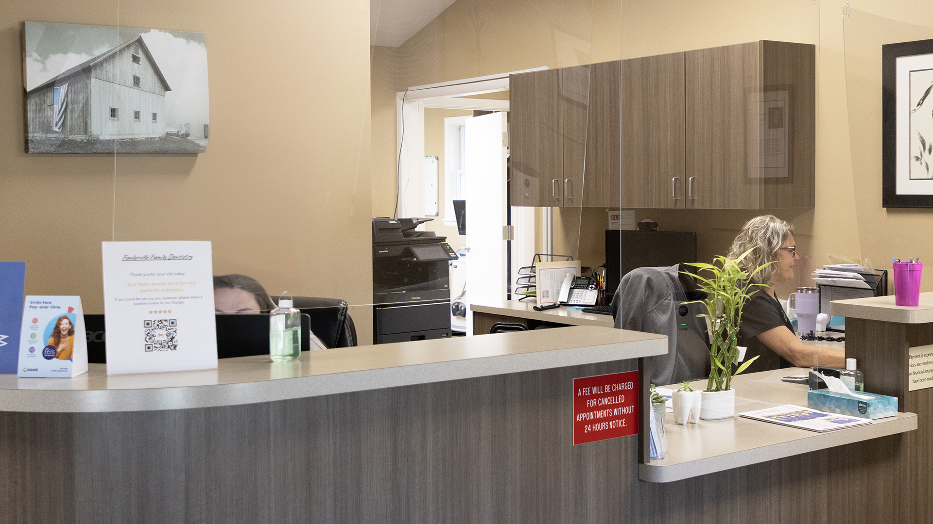 The image shows an interior view of a reception area in an office, featuring a front desk with a person sitting behind it and various items on the counter.