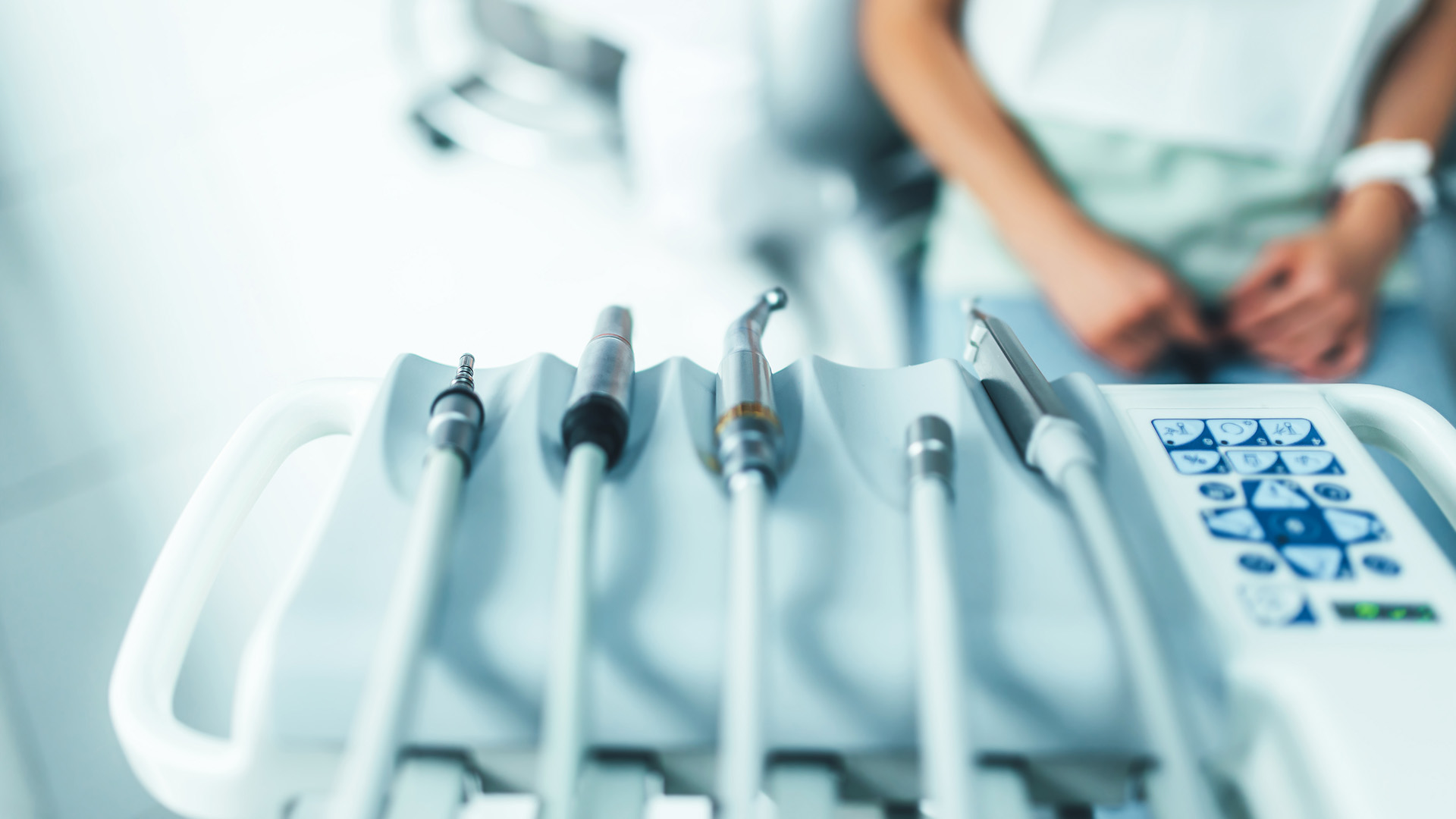 The image displays a medical setting with a person sitting in front of a tray containing various dental instruments, including a dental mirror and probes, and an electronic device that appears to be a dental X-ray machine.