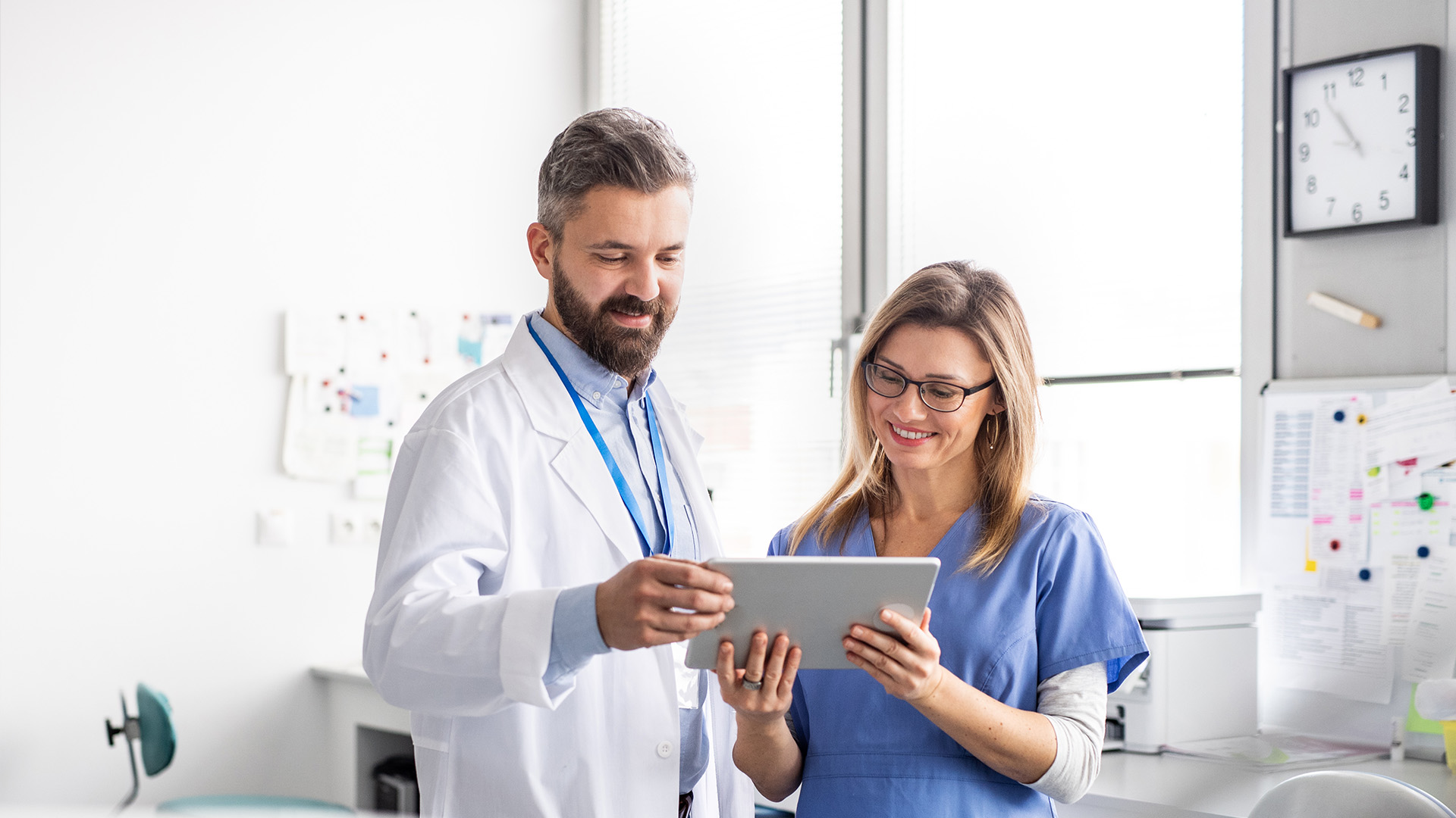 The image depicts a man and woman in a medical setting, with the man holding a tablet and both individuals looking at it.