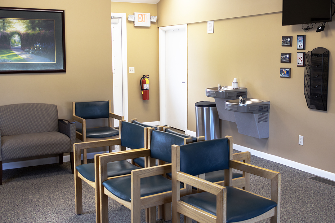 The image shows an interior space, likely a waiting area or lobby, with chairs arranged in rows and a sink counter with a faucet. There is a television mounted on the wall, and a framed picture hanging above it. The room has a neutral color palette with light-colored walls and flooring.