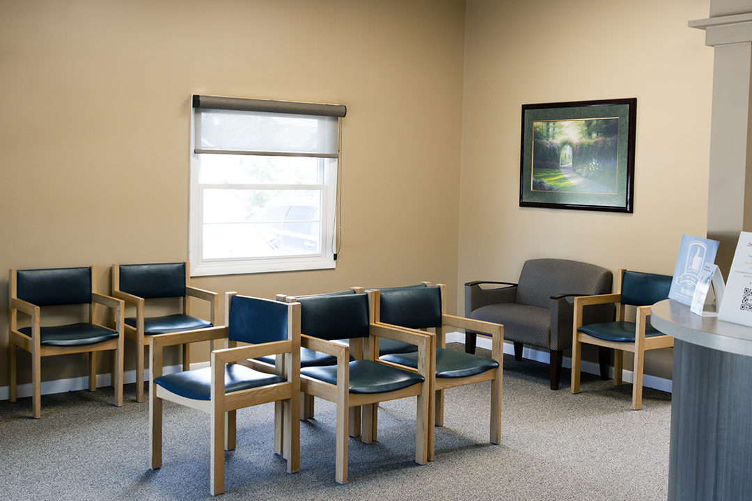 The image shows an interior view of a waiting room with several chairs arranged in rows, a wooden floor, and a window with white blinds partially drawn.