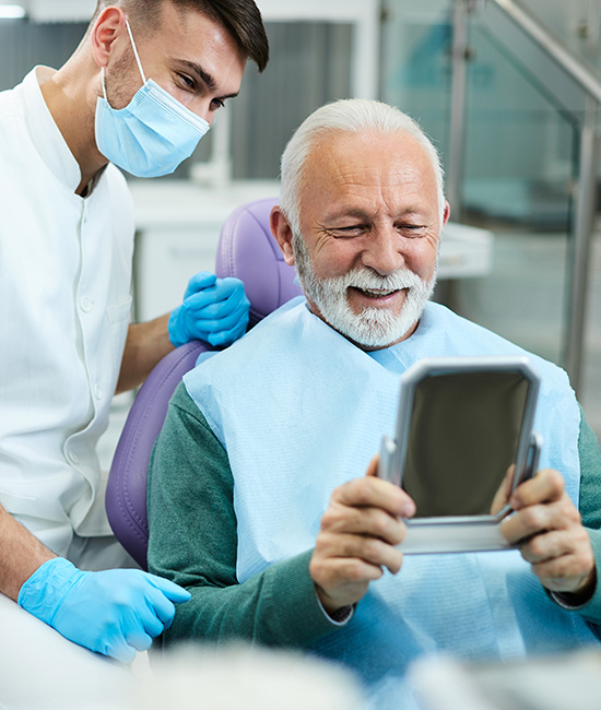 An elderly man with a beard is seated in a dental chair, looking at his reflection in a tablet device, while a dental professional stands beside him, smiling.