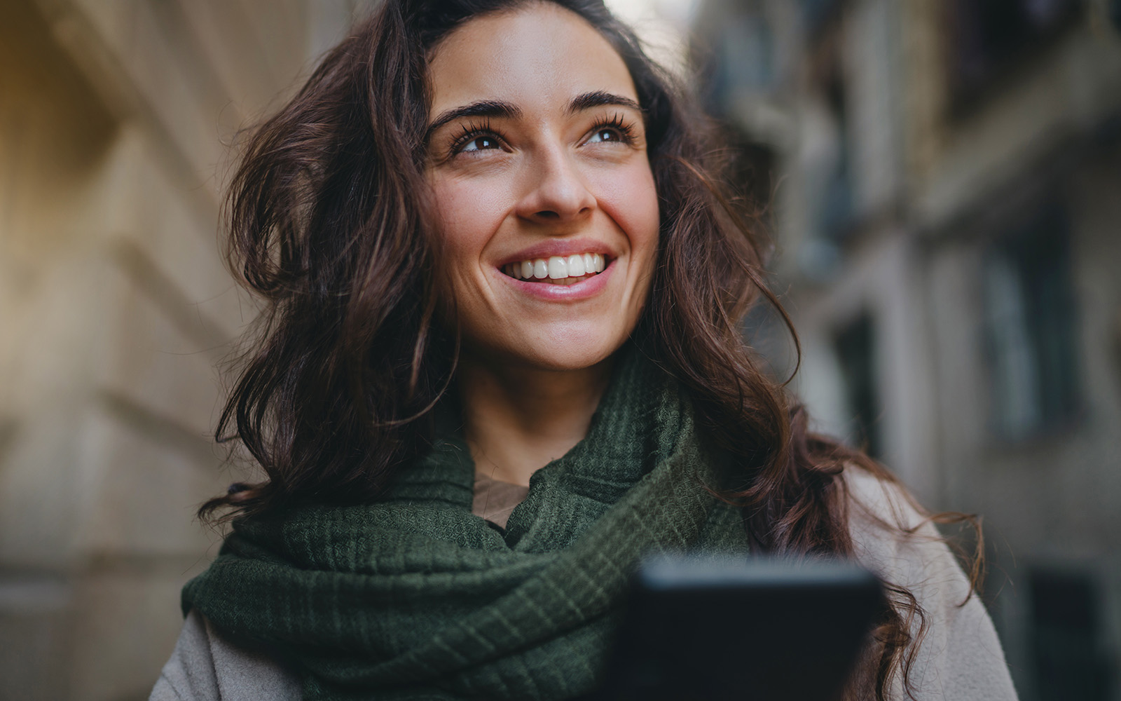 A woman is smiling and looking at her phone, standing on a city street.