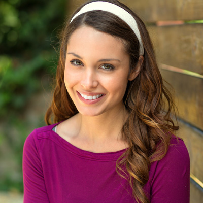 A smiling woman with long hair and a purple top, posing against a wooden fence.