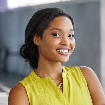 A young woman with a bright smile, wearing a yellow top and standing against a blue wall.