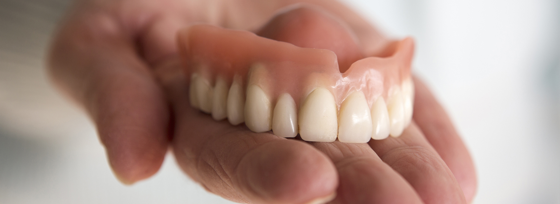 The image is a close-up photograph of a person s hand holding a set of artificial teeth with a blurred background, emphasizing the teeth.