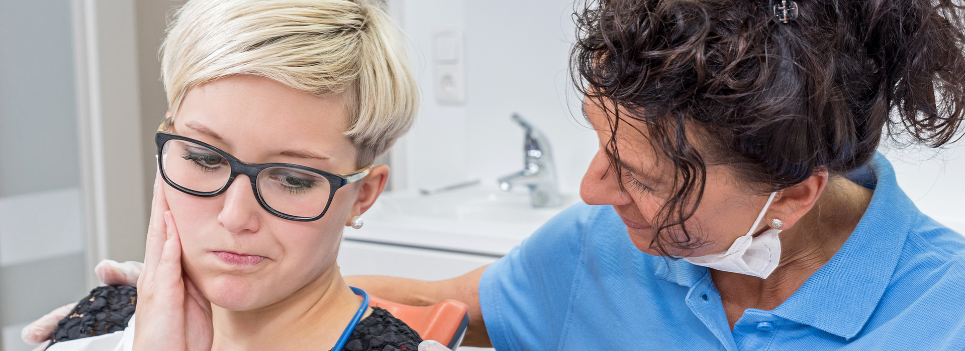 A woman with glasses is sitting in a dental chair, receiving treatment from a female dentist.