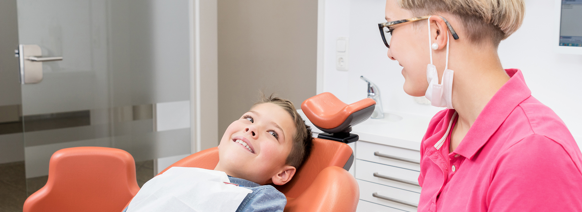 A dental hygienist is assisting a child in a dental chair, with the hygienist wearing a pink shirt and the child wearing a blue shirt.