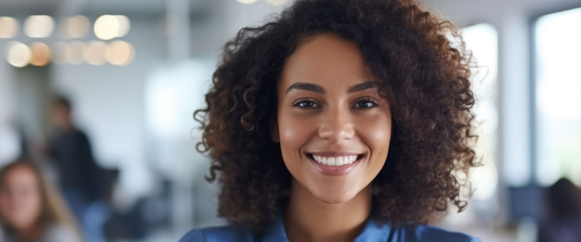 A smiling woman with curly hair, wearing a blue shirt, standing in an office environment.