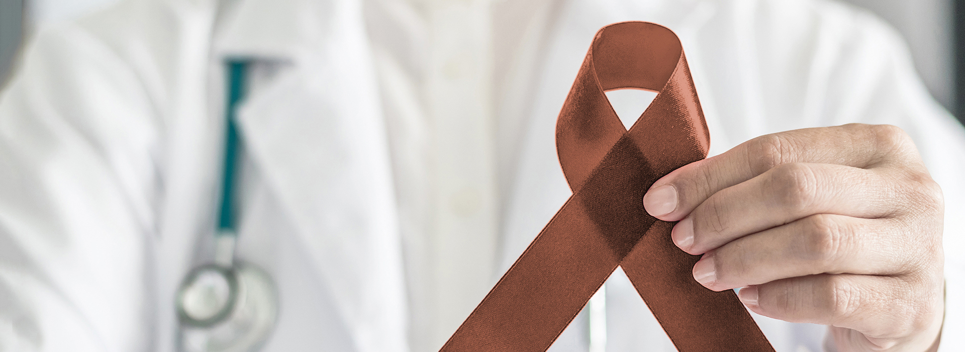 The image shows a person wearing a white lab coat holding up a brown ribbon with a red symbol on it, which is commonly associated with awareness for cancer.