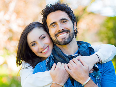 A man and a woman are hugging each other, smiling warmly, with the man wearing a scarf around his neck.