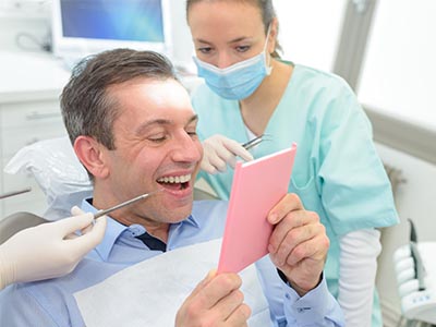 A man in a dental office, smiling and holding up a pink card with  20  written on it, surrounded by dental equipment and professionals.