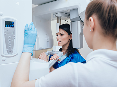 A woman in a blue lab coat is standing next to a large, modern medical imaging machine, which appears to be an MRI or CT scanner, while another person looks on.