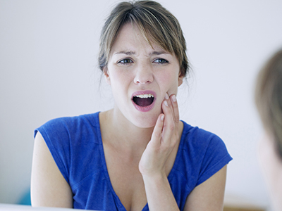 A woman with her mouth open, showing a toothbrush in her hand, and expressing surprise or concern while looking into the mirror.