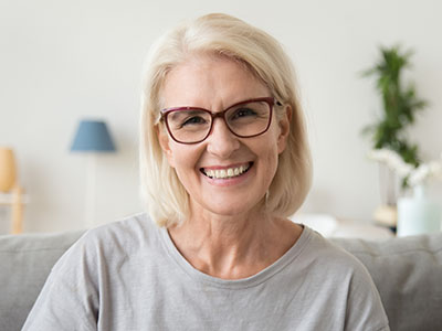 The image shows a middle-aged woman with blonde hair and glasses, smiling at the camera. She is wearing a light blue top and has short hair. The background is neutral and out of focus, suggesting an indoor setting.