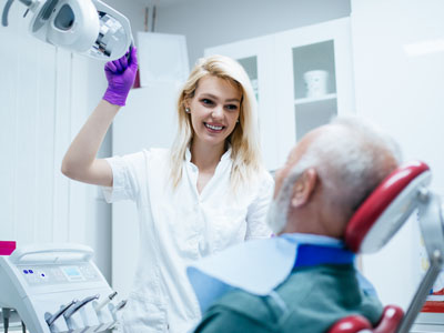 A woman in a white coat is assisting an elderly man with dental treatment, using a dental chair and equipment.